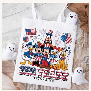Może przedstawiać: Biała torba z kolorową grafiką przedstawiającą Myszkę Miki, Myszkę Minnie, Kaczora Donalda, Daisy, Goofy'ego i Pluto w patriotycznych strojach. Napis 250 YEARS 1776-2026 znajduje się pod postaciami wraz z motywami flagi amerykańskiej.