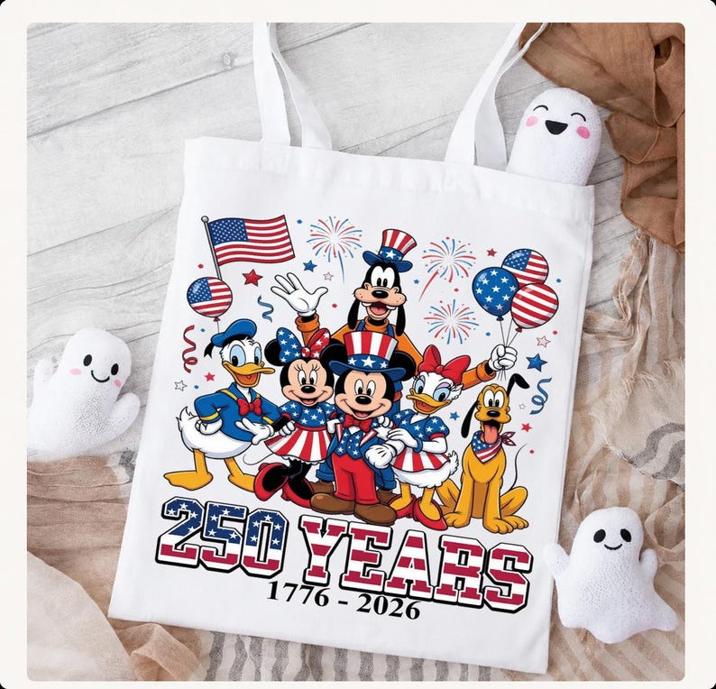 Może przedstawiać: Biała torba z kolorową grafiką przedstawiającą Myszkę Miki, Myszkę Minnie, Kaczora Donalda, Daisy, Goofy'ego i Pluto w patriotycznych strojach. Napis 250 YEARS 1776-2026 znajduje się pod postaciami wraz z motywami flagi amerykańskiej.