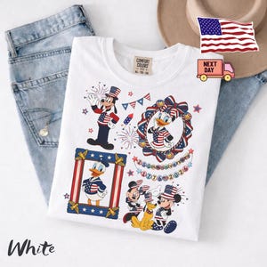 May include: A white t-shirt featuring a graphic print of Disney characters including Goofy, Daisy Duck, Donald Duck, Mickey Mouse, and Pluto wearing patriotic red, white, and blue outfits. The design includes fireworks, stars, and text reading 250th Anniversary 1776-2026.