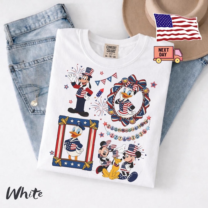 May include: A white t-shirt featuring a graphic print of Disney characters including Goofy, Daisy Duck, Donald Duck, Mickey Mouse, and Pluto wearing patriotic red, white, and blue outfits. The design includes fireworks, stars, and text reading 250th Anniversary 1776-2026.