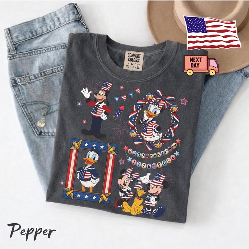 May include: A charcoal gray t-shirt featuring a patriotic graphic with Goofy, Donald Duck, Daisy Duck, Mickey Mouse, and Pluto wearing American flag-themed clothing. The design includes the text 250th Anniversary 1776-2026, stars, and fireworks.