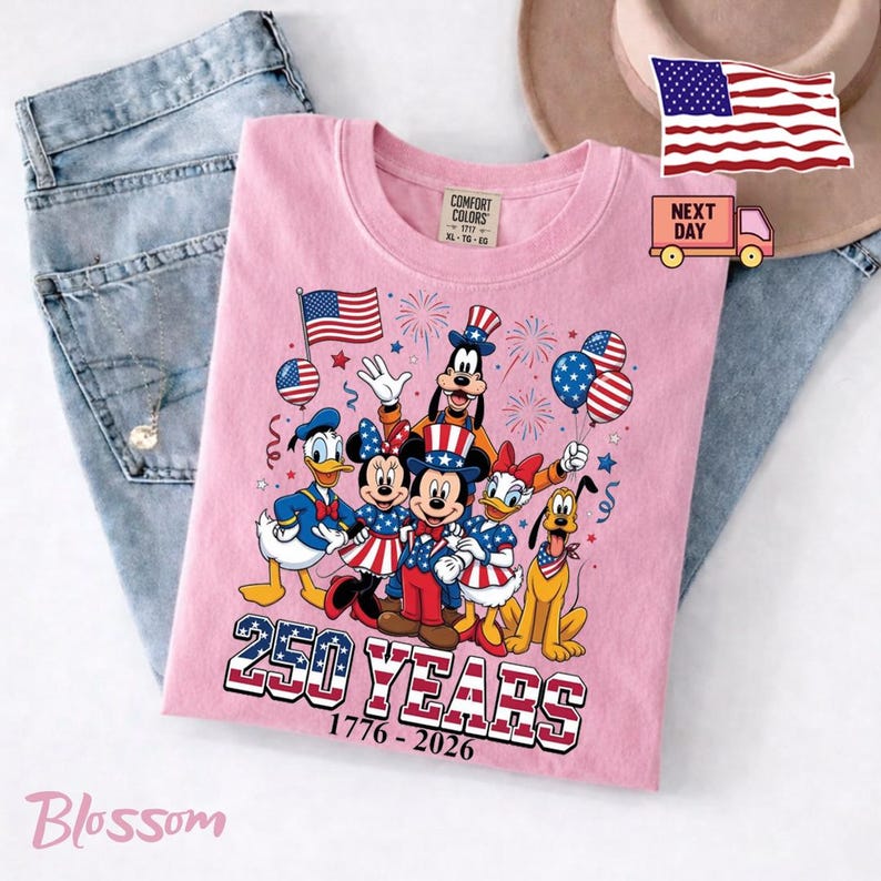 K&ouml;nnte beinhalten: Ein hellrosa T-Shirt mit einer Grafik von Disney-Figuren wie Mickey Maus, Minnie Maus, Donald Duck, Daisy Duck, Goofy und Pluto in patriotischer Kleidung. Der Text lautet 250 YEARS 1776-2026 mit amerikanischen Flaggenmustern.