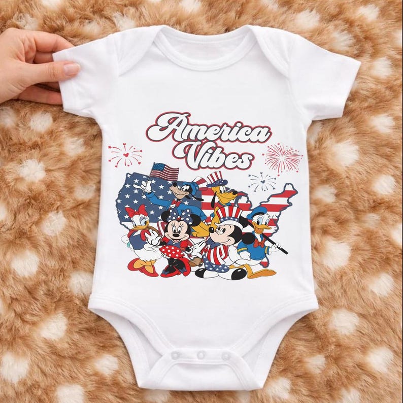 May include: A white short-sleeved baby bodysuit featuring a graphic print of Disney characters including Mickey Mouse, Minnie Mouse, Donald Duck, Daisy Duck, Goofy, and Pluto. The design includes a map of the USA and the text America Vibes.