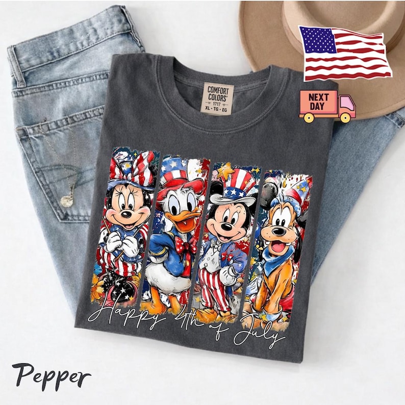 May include: A charcoal grey t-shirt featuring a graphic print of four cartoon characters wearing patriotic red, white, and blue outfits. The text "Happy 4th of July" is printed in white script below the characters. The shirt has a Comfort Colors label.