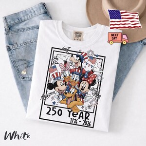 May include: A white t-shirt featuring a graphic of Disney characters including Mickey Mouse, Minnie Mouse, Donald Duck, Daisy Duck, Goofy, and Pluto wearing patriotic red, white, and blue attire. The text reads 250 Year 1776-2026 with character names.