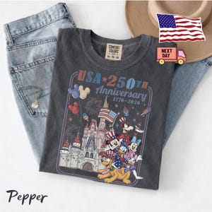 Peut inclure: Un t-shirt gris anthracite avec une impression graphique portant le texte USA 250TH Anniversary 1776-2026. Le motif comprend un ch&acirc;teau, des feux d'artifice, des ballons et des personnages de dessin anim&eacute; portant des chapeaux patriotiques.