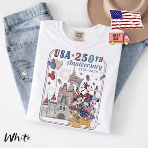 Peut inclure: Un t-shirt blanc avec une impression graphique repr&eacute;sentant un ch&acirc;teau, des feux d'artifice, des ballons et des personnages de dessin anim&eacute; portant des chapeaux patriotiques. Le texte indique USA 250TH Anniversary 1776-2026.