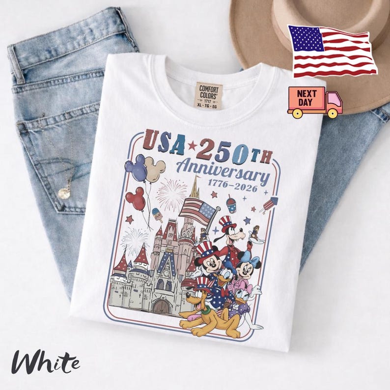 Peut inclure: Un t-shirt blanc avec une impression graphique repr&eacute;sentant un ch&acirc;teau, des feux d'artifice, des ballons et des personnages de dessin anim&eacute; portant des chapeaux patriotiques. Le texte indique USA 250TH Anniversary 1776-2026.