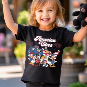 May include: A black short-sleeved t-shirt featuring a graphic print of cartoon characters in patriotic attire over a map of the United States. The text "America Vibes" is printed in white above the characters, accompanied by red firework illustrations.