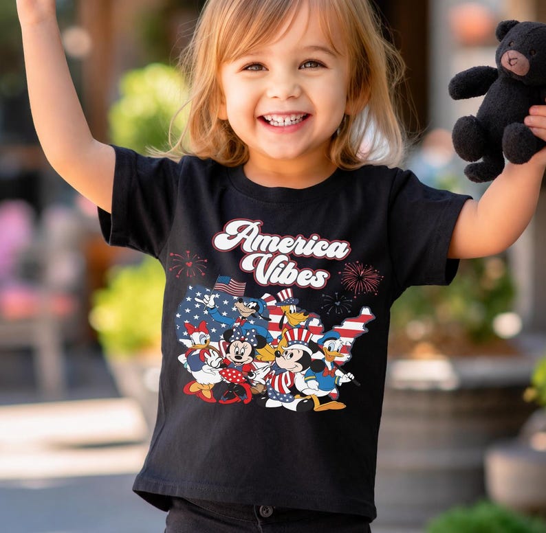 May include: A black short-sleeved t-shirt featuring a graphic print of cartoon characters in patriotic attire over a map of the United States. The text "America Vibes" is printed in white above the characters, accompanied by red firework illustrations.