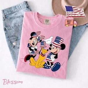 May include: A light pink t-shirt featuring a graphic of Minnie Mouse, Pluto, and Mickey Mouse wearing patriotic top hats and clothing with American flag patterns. The shirt has a Comfort Colors label at the neck and is displayed with blue jeans.