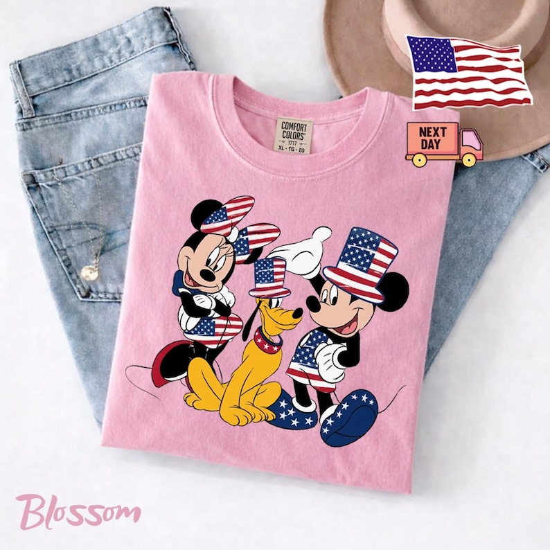 May include: A light pink t-shirt featuring a graphic of Minnie Mouse, Pluto, and Mickey Mouse wearing patriotic top hats and clothing with American flag patterns. The shirt has a Comfort Colors label at the neck and is displayed with blue jeans.