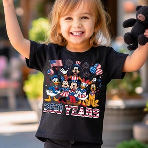 Może przedstawiać: Czarny t-shirt z kr&oacute;tkim rękawem z grafiką przedstawiającą postacie Disneya, w tym Myszkę Miki, Myszkę Minnie, Kaczora Donalda, Daisy, Goofy'ego i Pluto w patriotycznych strojach, z napisem 250 YEARS 1776-2026 oraz amerykańskimi flagami.