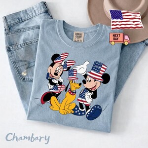 May include: A light blue crew neck t-shirt featuring a graphic of Minnie Mouse, Pluto, and Mickey Mouse wearing patriotic American flag-themed hats and clothing. The shirt has a Comfort Colors label and is paired with light blue denim jeans.