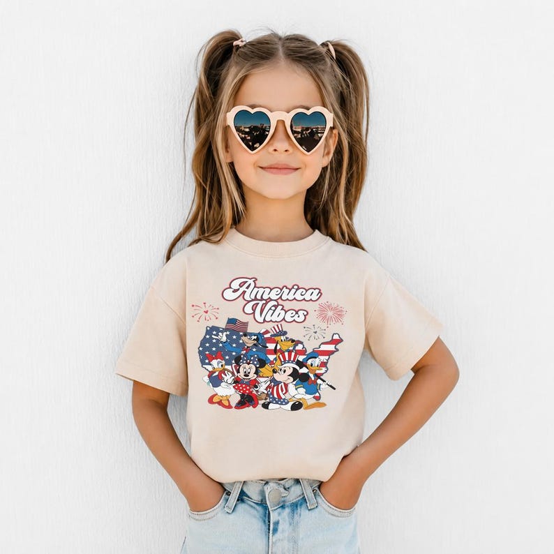 May include: A beige short-sleeve t-shirt featuring a graphic print of a map of the United States filled with a stars and stripes pattern, surrounded by Disney characters and the text America Vibes, worn with heart-shaped sunglasses and blue jeans.