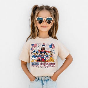 Może przedstawiać: Beżowy t-shirt z kr&oacute;tkim rękawem z grafiką postaci Disneya w patriotycznych strojach, z amerykańskimi flagami, balonami i fajerwerkami. Napis głosi 250 YEARS 1776-2026 w czerwono-biało-niebieskiej czcionce w paski.