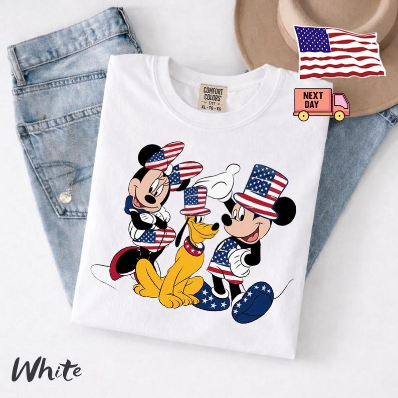 May include: A white t-shirt featuring a graphic of Minnie Mouse, Pluto, and Mickey Mouse wearing patriotic top hats and clothing with American flag patterns. The shirt has a Comfort Colors label at the neck and is displayed on blue denim fabric.