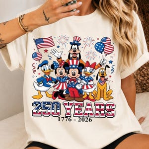 Może przedstawiać: Kremowy t-shirt z grafiką przedstawiającą postacie Disneya, w tym Myszkę Miki, Myszkę Minnie, Kaczora Donalda, Daisy, Goofy'ego i Pluto w patriotycznych strojach. Napis głosi 250 YEARS 1776 - 2026, w tle widać fajerwerki.