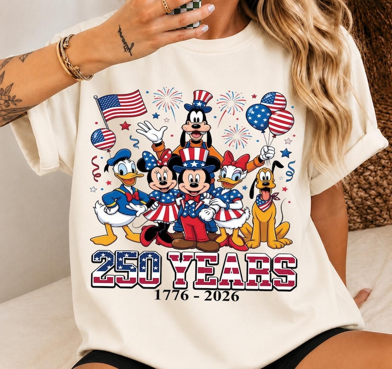 Może przedstawiać: Kremowy t-shirt z grafiką przedstawiającą postacie Disneya, w tym Myszkę Miki, Myszkę Minnie, Kaczora Donalda, Daisy, Goofy'ego i Pluto w patriotycznych strojach. Napis głosi 250 YEARS 1776 - 2026, w tle widać fajerwerki.