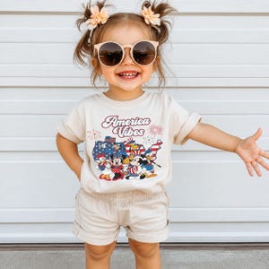 May include: A cream-colored short-sleeve t-shirt featuring a graphic of cartoon characters in front of a red, white, and blue map of the United States with the text America Vibes and fireworks. The shirt is paired with matching cream-colored shorts.