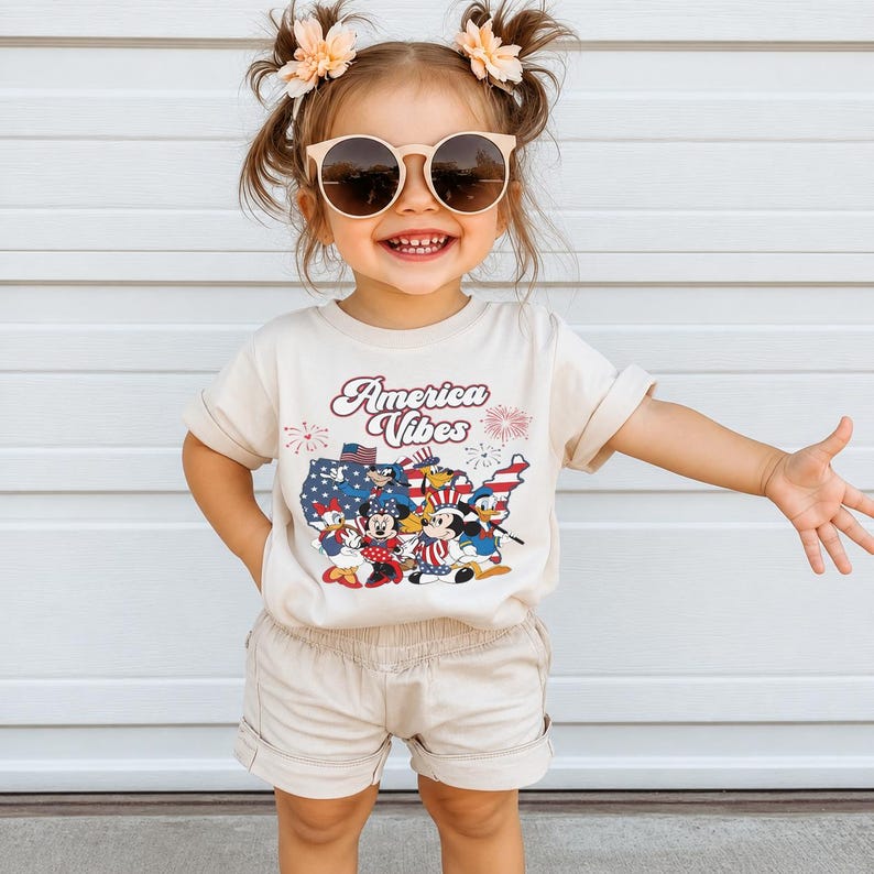 May include: A cream-colored short-sleeve t-shirt featuring a graphic of cartoon characters in front of a red, white, and blue map of the United States with the text America Vibes and fireworks. The shirt is paired with matching cream-colored shorts.
