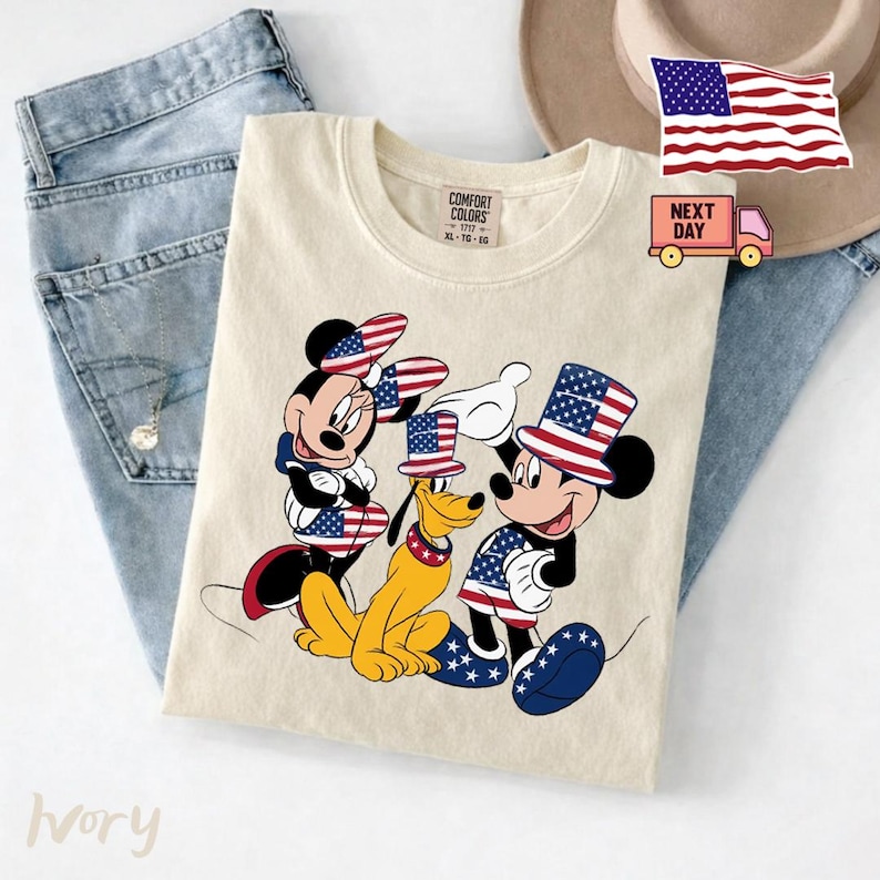 May include: An ivory t-shirt featuring a graphic of Minnie Mouse, Pluto, and Mickey Mouse wearing patriotic American flag-themed hats and clothing. The shirt has a Comfort Colors label at the neck and is displayed alongside blue denim jeans.