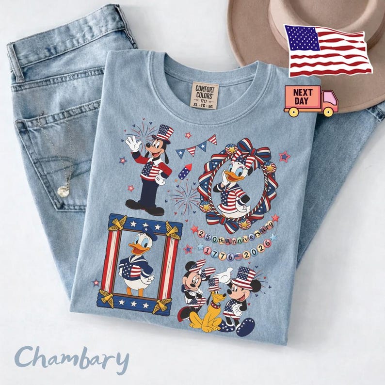 May include: A light blue t-shirt featuring a graphic print of cartoon characters wearing patriotic red, white, and blue clothing. The design includes fireworks, stars, and text reading 250th anniversary 1776-2026. The shirt has a Comfort Colors brand label.