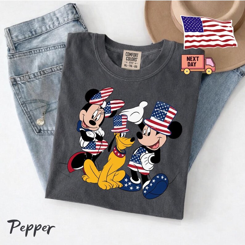 May include: A charcoal gray t-shirt featuring a graphic of Minnie Mouse, Pluto, and Mickey Mouse wearing patriotic American flag-themed hats and clothing. The shirt has a Comfort Colors label at the neck and the text Pepper is printed below.