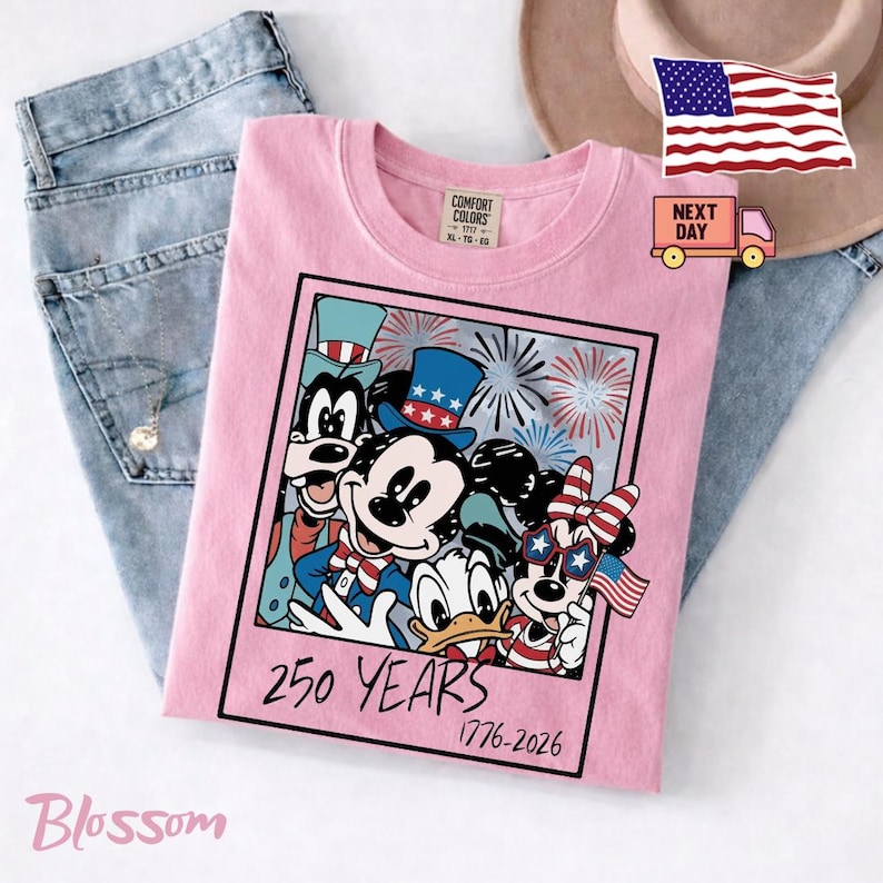 May include: A light pink t-shirt featuring a graphic of cartoon characters wearing patriotic hats and accessories. The design includes fireworks and the text 250 YEARS 1776-2026. The shirt is layered over blue denim jeans and a tan hat.