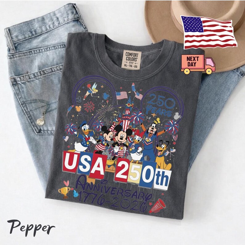 May include: A charcoal gray t-shirt featuring a graphic of Disney characters including Mickey Mouse, Minnie Mouse, Donald Duck, Daisy Duck, Goofy, Pluto, and Tinker Bell. The text reads USA 250th Anniversary 1776-2026 with fireworks and a castle.