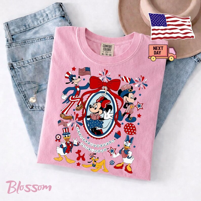 Może przedstawiać: Jasnor&oacute;żowy t-shirt z nadrukiem graficznym przedstawiającym postacie Disneya, w tym Myszkę Miki, Minnie, Goofy'ego, Kaczora Donalda, Daisy i Pluto w patriotycznych czerwono-biało-niebieskich strojach. Wz&oacute;r zawiera dużą czerwoną kokardę, fajerwerki oraz napis 250th Anniversary 1776-2026.