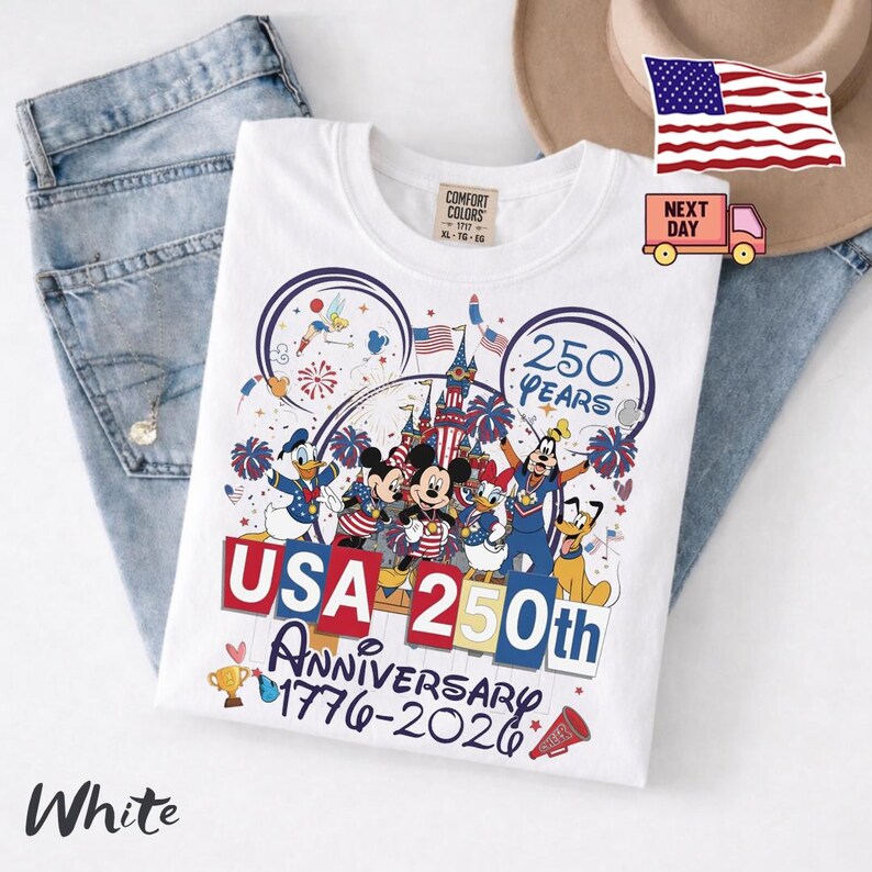 May include: A white t-shirt featuring a graphic of Disney characters including Mickey Mouse, Donald Duck, and Goofy. The design includes the text USA 250th ANNIVERSARY 1776-2026, fireworks, a castle, and American flags. The shirt is displayed on blue denim.