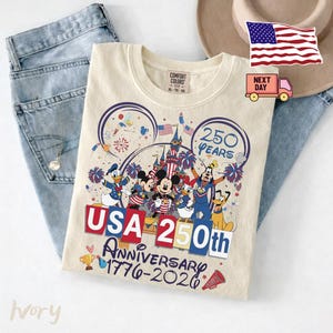 May include: An ivory t-shirt featuring a graphic of Disney characters including Mickey Mouse, Donald Duck, and Goofy in front of a castle. The text reads 250 YEARS, USA 250th ANNIVERSARY 1776-2026, with fireworks, American flags, and a trophy illustration.