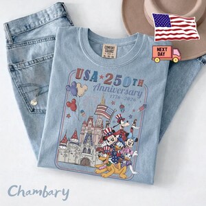 Peut inclure: Un t-shirt bleu clair avec un motif repr&eacute;sentant un ch&acirc;teau, des feux d'artifice et des personnages de dessin anim&eacute; portant des chapeaux patriotiques. Le texte indique USA 250TH Anniversary 1776-2026. Accompagn&eacute; d'un jean bleu clair et d'un chapeau beige.