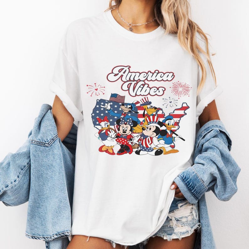 May include: A white t-shirt featuring a graphic print of cartoon characters including Mickey Mouse, Minnie Mouse, Donald Duck, Daisy Duck, and Pluto. The design includes a map of the United States, fireworks, and the text America Vibes in red.