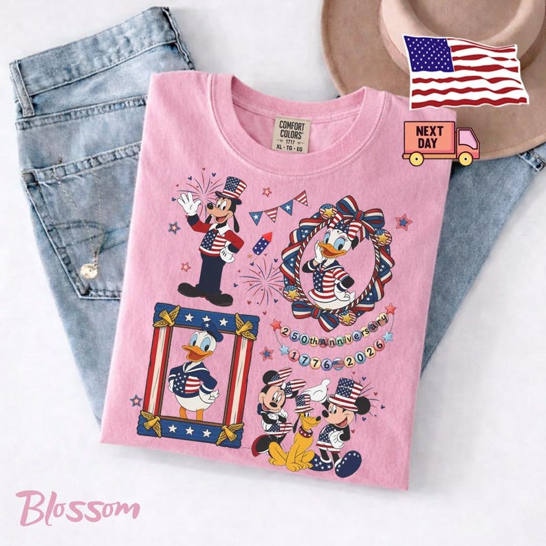 May include: A light pink t-shirt featuring a graphic print of Disney characters including Goofy, Daisy Duck, Donald Duck, Mickey Mouse, and Pluto wearing patriotic red, white, and blue clothing. The text reads 250th Anniversary 1776-2026.