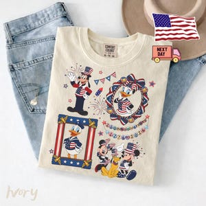 May include: An ivory t-shirt featuring a graphic print of Goofy, Daisy Duck, Donald Duck, Mickey Mouse, and Pluto wearing patriotic American flag-themed clothing. The design includes fireworks, bunting, and text reading 250th Anniversary 1776-2026.