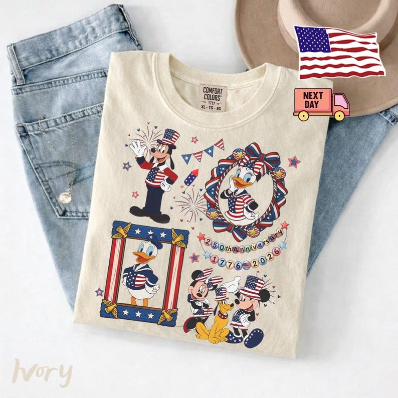 May include: An ivory t-shirt featuring a graphic print of Goofy, Daisy Duck, Donald Duck, Mickey Mouse, and Pluto wearing patriotic American flag-themed clothing. The design includes fireworks, bunting, and text reading 250th Anniversary 1776-2026.