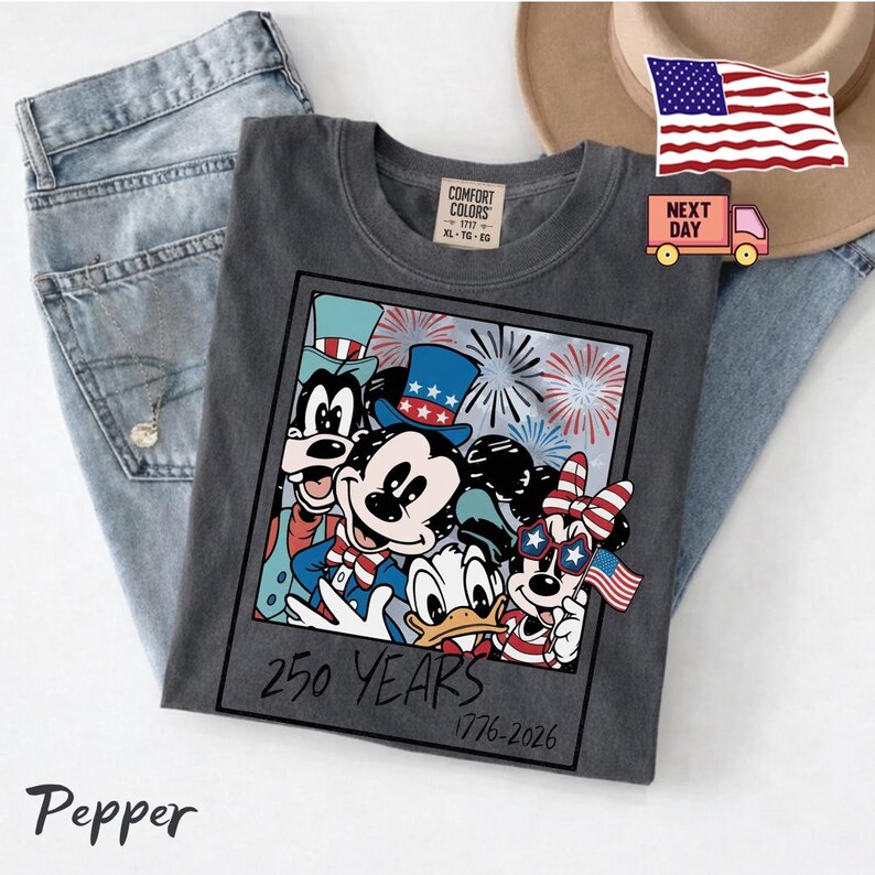 May include: A charcoal grey t-shirt featuring a graphic of Mickey Mouse, Minnie Mouse, Donald Duck, and Goofy wearing patriotic hats and accessories. The design includes fireworks and the text 250 YEARS 1776-2026. The shirt has a Comfort Colors label.