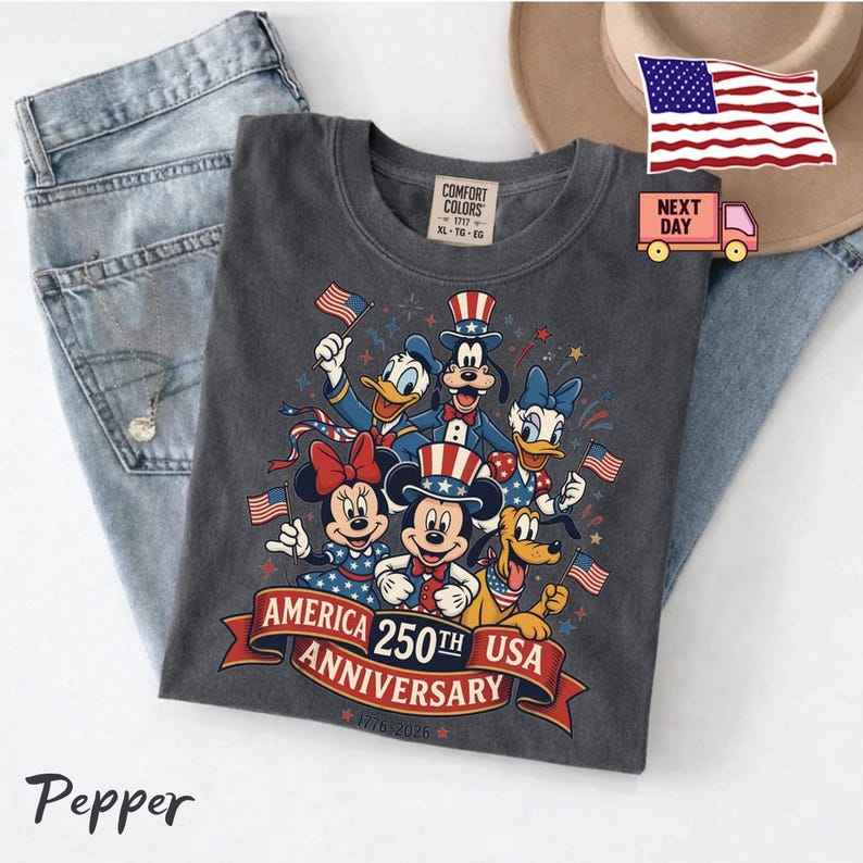 May include: A charcoal grey t-shirt featuring a graphic of Mickey Mouse, Minnie Mouse, Donald Duck, Daisy Duck, Goofy, and Pluto wearing patriotic attire. The text reads America 250th Anniversary 1776-2026. The shirt is displayed with blue jeans.