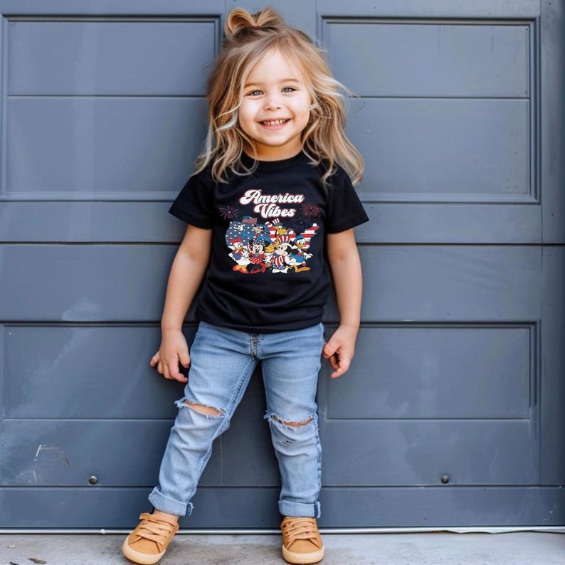 May include: A black short-sleeved t-shirt featuring the text America Vibes in white script above a graphic of the United States map filled with stars and stripes, accompanied by cartoon characters wearing patriotic red, white, and blue clothing.