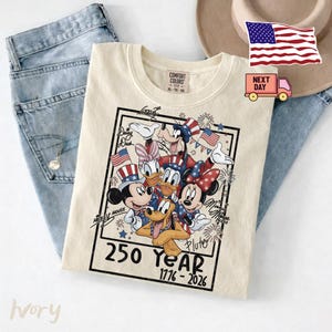 May include: An ivory t-shirt featuring a graphic of Disney characters including Mickey Mouse, Minnie Mouse, Donald Duck, Daisy Duck, Goofy, and Pluto wearing patriotic hats. The text reads 250 YEAR 1776-2026 with signatures and fireworks.