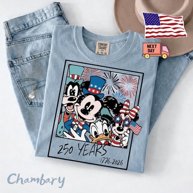 May include: A light blue t-shirt featuring a graphic of cartoon characters wearing patriotic hats and accessories, with fireworks in the background. The text reads 250 YEARS 1776-2026. The shirt has a Comfort Colors label at the inner neckline.