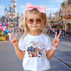 May include: A white short-sleeved t-shirt featuring a graphic of the United States map filled with stars and stripes, surrounded by cartoon characters and the text America Vibes. The design includes fireworks and patriotic colors of red, white, and blue.
