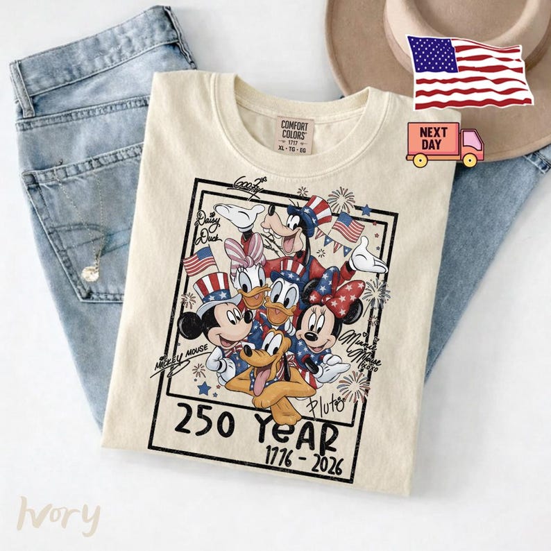 May include: An ivory t-shirt featuring a graphic of Disney characters including Mickey Mouse, Minnie Mouse, Donald Duck, Daisy Duck, Goofy, and Pluto wearing patriotic hats. The text reads 250 YEAR 1776-2026 with signatures and fireworks.