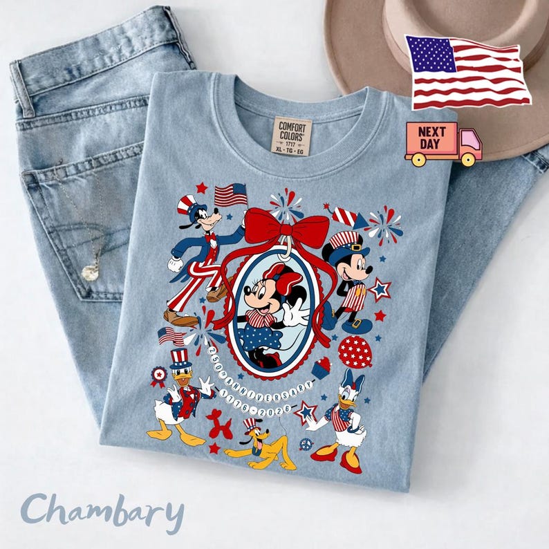 Może przedstawiać: Jasnoniebieski t-shirt z grafiką przedstawiającą postacie Disneya, w tym Myszkę Miki, Minnie, Kaczora Donalda, Goofy'ego i Pluto w patriotycznych czerwono-biało-niebieskich strojach. Wz&oacute;r zawiera dużą czerwoną kokardę, fajerwerki i napis 25th Anniversary 1996-2021.