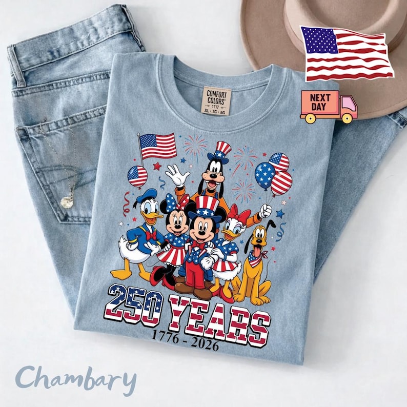 K&ouml;nnte beinhalten: Ein hellblaues T-Shirt mit einer Grafik von Disney-Figuren wie Mickey Maus, Minnie Maus, Donald Duck, Daisy Duck, Goofy und Pluto in patriotischer Kleidung. Der Text lautet 250 YEARS 1776-2026 mit US-Flaggenmustern.