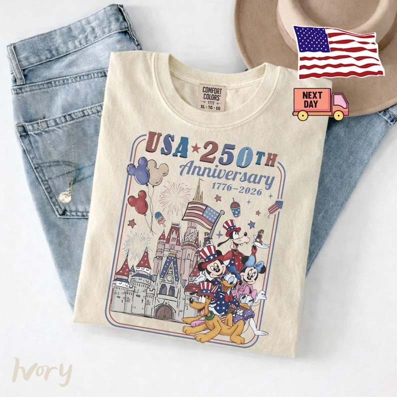 Peut inclure: Un t-shirt ivoire avec un motif repr&eacute;sentant un ch&acirc;teau, des feux d'artifice et des personnages de dessin anim&eacute; portant des chapeaux patriotiques. Le texte indique USA 250TH Anniversary 1776-2026. Le design inclut des ballons, un drapeau et des &eacute;toiles.