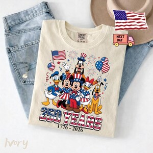 K&ouml;nnte beinhalten: Ein elfenbeinfarbenes T-Shirt mit einer Grafik von Zeichentrickfiguren in patriotischen rot-wei&szlig;-blauen Outfits. Das Design zeigt Feuerwerk, Luftballons und den Text 250 YEARS 1776-2026. Das Shirt hat ein Comfort Colors Etikett am Kragen.