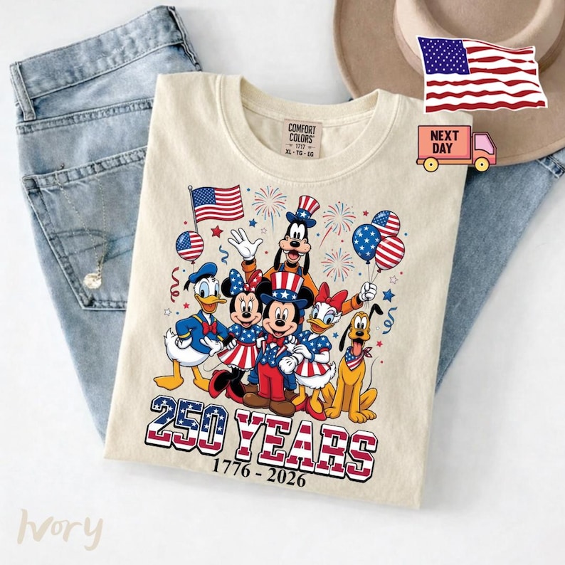 K&ouml;nnte beinhalten: Ein elfenbeinfarbenes T-Shirt mit einer Grafik von Zeichentrickfiguren in patriotischen rot-wei&szlig;-blauen Outfits. Das Design zeigt Feuerwerk, Luftballons und den Text 250 YEARS 1776-2026. Das Shirt hat ein Comfort Colors Etikett am Kragen.